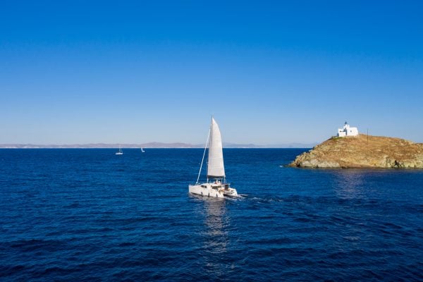 Sailing. Sailboat catamaran with white sails, rippled sea background, Lighthouse on a cape. Greece, Kea Tzia island. Summer holidays in Aegean sea. Aerial drone view